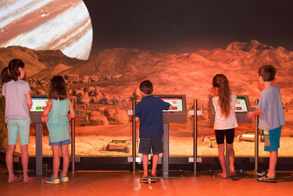 Five kids standing at touch screen kiosks, controlling small rovers inside of an enclosure with a landscape of the Io in the background.